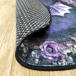Puede incluir: Una alfombra redonda de color negro y morado con un dise&ntilde;o floral y un murci&eacute;lago negro con alas moradas. La alfombra tiene un borde negro y un respaldo a cuadros.