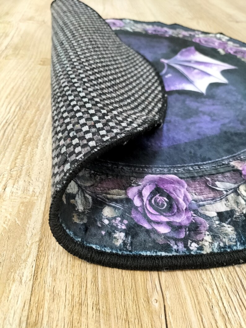 Puede incluir: Una alfombra redonda de color negro y morado con un dise&ntilde;o floral y un murci&eacute;lago negro con alas moradas. La alfombra tiene un borde negro y un respaldo a cuadros.