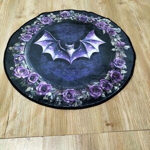 Puede incluir: Una alfombra redonda de color negro y morado con un dise&ntilde;o de murci&eacute;lago en el centro. El murci&eacute;lago est&aacute; rodeado de una corona de rosas moradas.