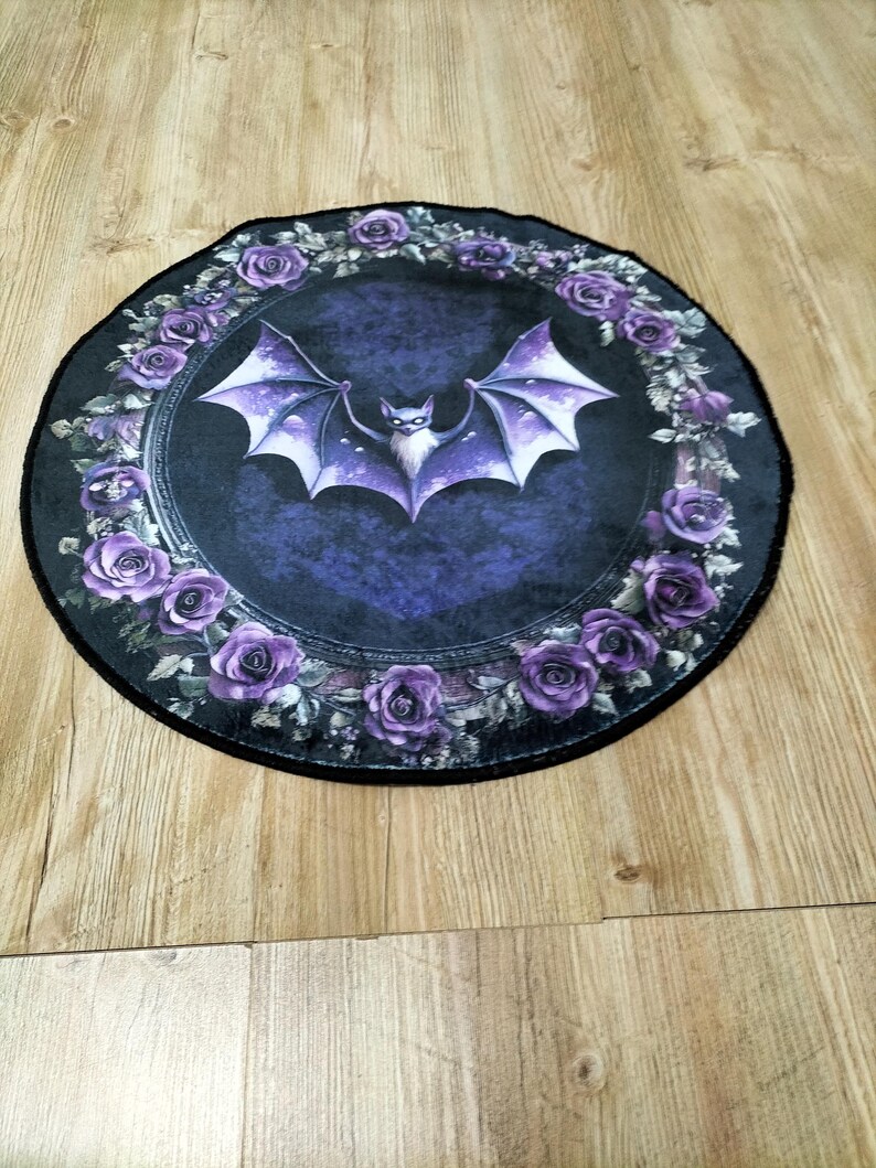 Puede incluir: Una alfombra redonda de color negro y morado con un dise&ntilde;o de murci&eacute;lago en el centro. El murci&eacute;lago est&aacute; rodeado de una corona de rosas moradas.