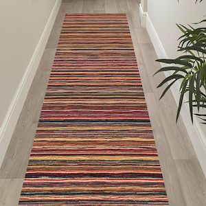 Op de afbeelding: Een lange, rechthoekige loper met een dicht patroon van horizontale strepen in diverse kleuren, waaronder tinten rood, oranje, geel, blauw en donkere kleuren. Het kleed heeft een getextureerd oppervlak en ligt op de vloer.