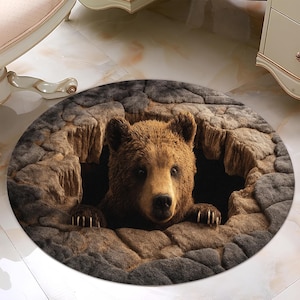 Op de afbeelding: Een ronde vloerkleed met een realistische bruine beer die uit een grot komt. Het vloerkleed heeft een getextureerde, steenachtige rand in grijze en bruine tinten, waardoor een 3D-effect ontstaat. De vacht van de beer is gedetailleerd en zijn poten zijn zichtbaar.