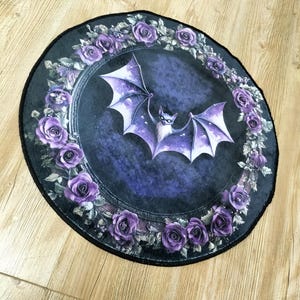 Puede incluir: Una alfombra redonda de color negro y morado con un dise&ntilde;o de murci&eacute;lago en el centro. El murci&eacute;lago est&aacute; rodeado de una corona de rosas moradas.