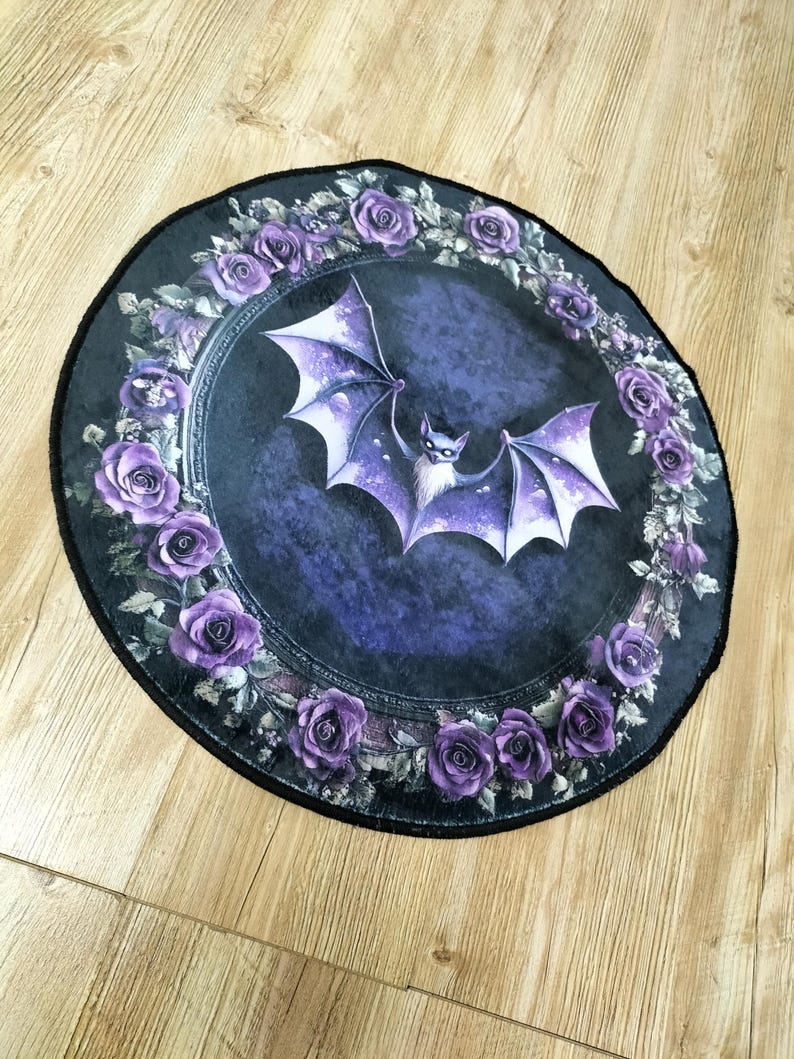 Puede incluir: Una alfombra redonda de color negro y morado con un dise&ntilde;o de murci&eacute;lago en el centro. El murci&eacute;lago est&aacute; rodeado de una corona de rosas moradas.