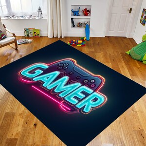 May include: A rectangular rug with a neon-style design featuring the word "GAMER" in bright blue, outlined in pink, and a stylized game controller. The rug is black with a gradient effect.