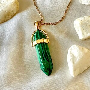 May include: A gold chain necklace with a green malachite crystal pendant. The pendant is a long, pointed crystal with a gold band around the top.