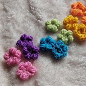 Puede incluir: Una colección de pequeñas aplicaciones de flores tejidas a ganchillo dispuestas en un espectro de colores que incluye rojo, naranja, amarillo, verde claro, azul, morado y rosa. Cada flor presenta un diseño de cinco pétalos con textura.