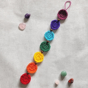 Może przedstawiać: Girlanda zrobiona na szydełku w kolorach tęczy, z siedmioma okrągłymi elementami, każdy w innym kolorze tęczy. Girlanda jest ozdobiona brązowymi koralikami i ma pętelkę na górze do zawieszenia.