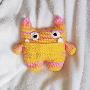 May include: A handmade crochet monster plush toy featuring horizontal stripes in vibrant yellow, orange, and pink. The creature has two pointed ears, two round white eyes with black pupils, two small white teeth, and a large yellow pocket on its front.