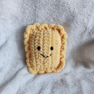 May include: A handmade, plush, yellow crocheted ravioli with a smiling face. The ravioli has a textured surface and a scalloped edge. The eyes are small black dots, and the mouth is a curved black line. The ravioli is laying on a white, fluffy surface.