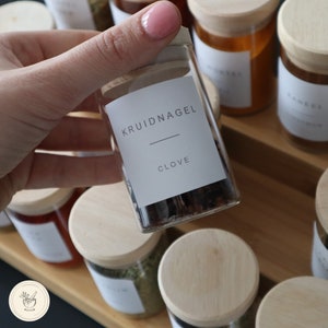 Op de afbeelding: Een glazen pot met een houten deksel gevuld met kruidnagels. De pot heeft een wit etiket met de tekst "KRUIDNAGEL" en "CLOVE" erop.