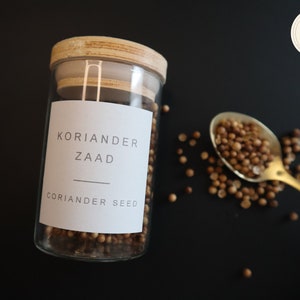Op de afbeelding: Een glazen pot met een houten deksel gevuld met korianderzaad. De pot heeft een wit etiket met de tekst "KORIANDER ZAAD" en "CORIANDER SEED". Een lepel met korianderzaad ligt op een zwart oppervlak.