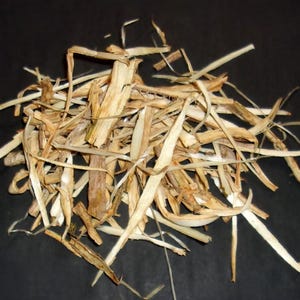 May include: A pile of dried, light brown plant material with long, thin, and irregular shapes. The pieces vary in color from light tan to darker brown, with some having a slightly curved appearance. The background is a solid black.