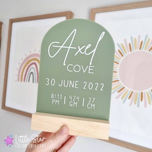 May include: A sage green acrylic sign with rounded edges and a wooden stand. The sign reads "Axel Cove" and includes the date "30 June 2022" and the time "8:11 PM". The sign also includes the measurements "424 g" and "27 cm".