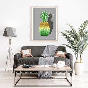 Könnte beinhalten: Ein gerahmter Druck eines gelben und grünen Ananasses mit einem Schatten auf grauem Hintergrund. Der Druck hängt an einer weißen Wand über einem grauen Ledersofa mit einer Wolldecke und zwei Kissen. Ein Holz-Couchtisch mit einer weißen Keramiktasse und einer kleinen Schale steht vor dem Sofa. Eine Topfpalme steht rechts im Bild.