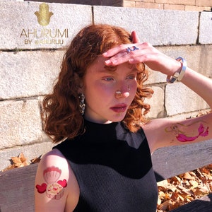 Puede incluir: Una persona con cabello rojo que lleva una camisa negra de cuello alto y tiene varios tatuajes temporales en los brazos. Los tatuajes son de una sirena, una concha y una corona. El texto "AHURUMI BY @AHURUU" es visible en la esquina superior izquierda de la imagen.