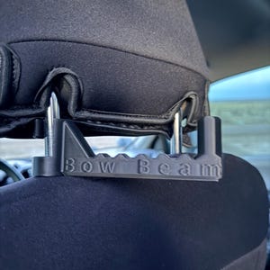 May include: A black plastic car headrest mount with the text "Bow Beam" is attached to a black car seat headrest. The mount has a series of slots and a triangular support.