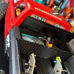 May include: A red toy truck with a black bed and a battery compartment. The truck has a "FORD" logo and Route 66 stickers. The bed has a diamond plate pattern and contains a green battery and wiring.