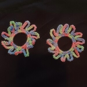May include: Two handmade crochet hair scrunchies in a flower design. The scrunchies feature a circular center with colorful petals in shades of pink, blue, green, and yellow. The background is a solid black.