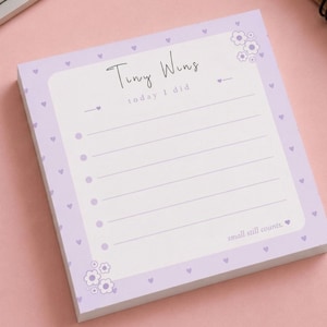 May include: A square notepad with a light purple border adorned with small hearts and floral accents. The notepad features the text "Tiny Wins today I did" and lined spaces for notes. The phrase "small still counts" is at the bottom.