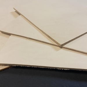 May include: Close-up of several stacked, light tan leather sheets. The edges of the leather are visible, showing the thickness and texture. The sheets are arranged at different angles, creating a layered effect.