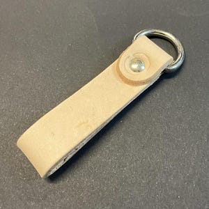 May include: A light tan leather keyring with a silver metal ring. The rectangular leather strap is secured with a silver rivet. The keyring is designed for holding keys.