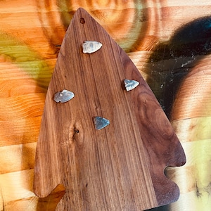 May include: A wooden cutting board with a unique shape, featuring a rich brown color and visible wood grain. The board is adorned with four small, arrowhead-shaped stones. A hole is drilled at the top for hanging.