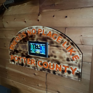 May include: Wooden sign with the text "THERE'S NO PLACE LIKE POTTER COUNTY" in orange lettering. The sign has a built-in digital weather station displaying temperature and other data. The sign is mounted on a wooden wall.