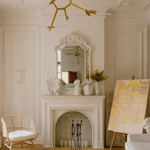 May include: A white living room with a fireplace, a gold chandelier, a mirror, and a white sofa. There is a sculpture of three figures in front of the fireplace.