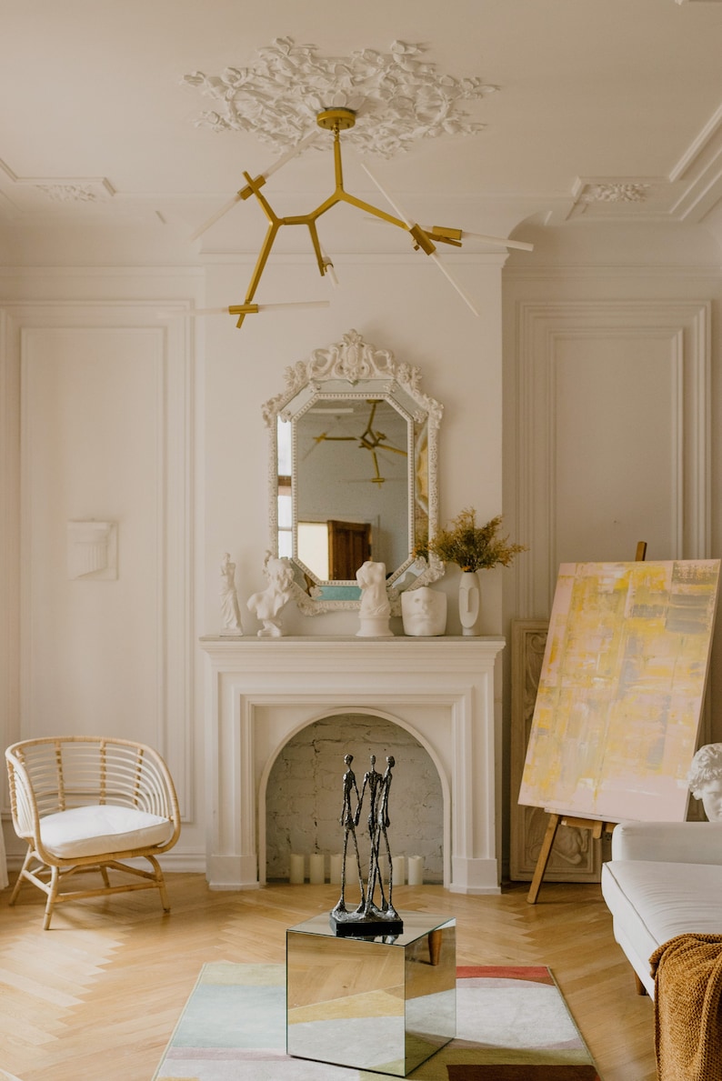 May include: A white living room with a fireplace, a gold chandelier, a mirror, and a white sofa. There is a sculpture of three figures in front of the fireplace.