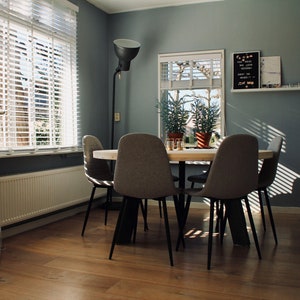 May include: A dining room with a wooden table and four grey chairs. The table is set with glasses and a white tablecloth. There is a large window with white blinds and a floor lamp in the corner. The walls are painted a light grey.
