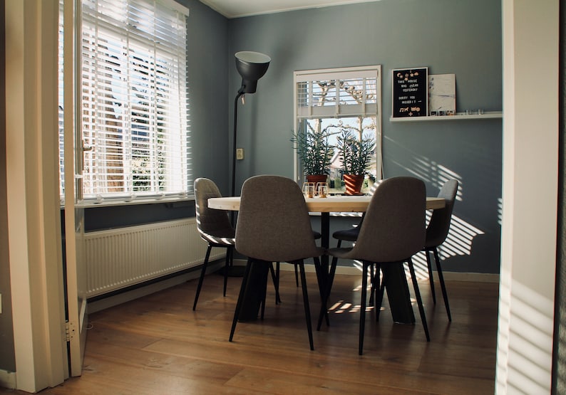 May include: A dining room with a wooden table and four grey chairs. The table is set with glasses and a white tablecloth. There is a large window with white blinds and a floor lamp in the corner. The walls are painted a light grey.