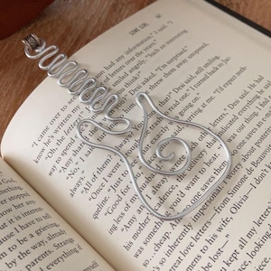 May include: A silver wire bookmark shaped like a guitar. The bookmark is laying on a book with the text "Gone Girl" and a page number of 167. The text on the page reads "I came over to see if he's written my way to spend the letters!"