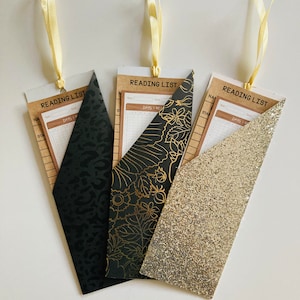 May include: Three reading list bookmarks with different patterned covers. The covers are black with gold floral designs, black with gold leopard print, and gold glitter. Each bookmark has a checklist for tracking reading progress.