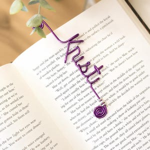 Puede incluir: Un marcapáginas de alambre morado con la palabra "Kristi" en cursiva, un extremo en espiral y una pequeña cuenta plateada. El marcapáginas está sobre una página de libro abierta y tiene un elemento decorativo de hoja verde en la parte superior.