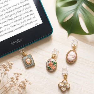 May include: Four decorative gold-toned metal charms with clear plastic clips, including a rectangular charm with an opal-like center, an oval floral charm, an oval cameo charm with a pink background, and a four-leaf clover charm with white pearls.