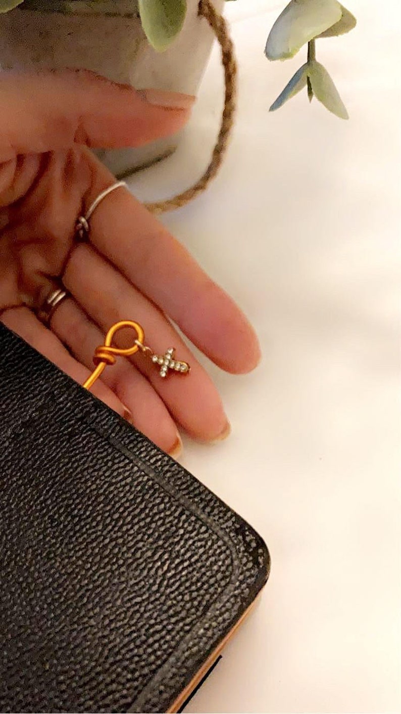 May include: A gold-colored bookmark with a small, sparkly cross charm. The bookmark is tucked into the corner of a black leather-bound book.