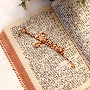 May include: A gold-colored wire bookmark shaped like the word "Jesus" is placed in an open Bible. The bookmark is resting on the page with the text "EZRA, 8."