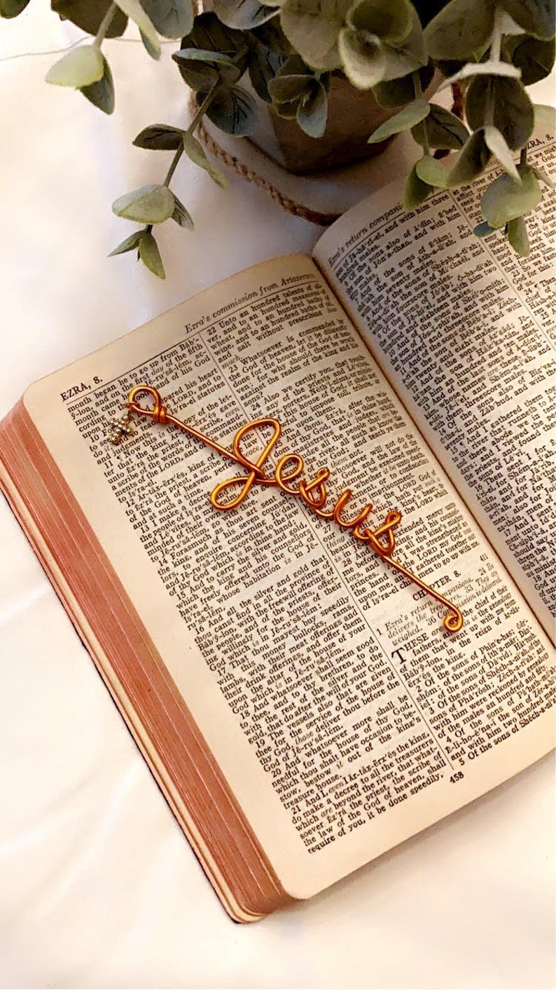 May include: A gold-colored wire bookmark shaped like the word "Jesus" is placed in an open Bible. The bookmark is resting on the page with the text "EZRA, 8."