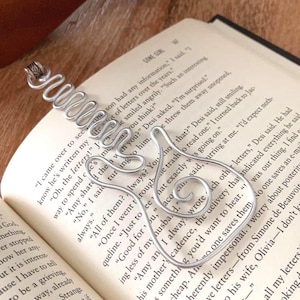 May include: A silver-colored metal bookmark shaped like a treble clef, featuring a coiled wire design at the top. The bookmark is resting on an open book with printed text, with a portion of a wooden stringed instrument visible at the top.