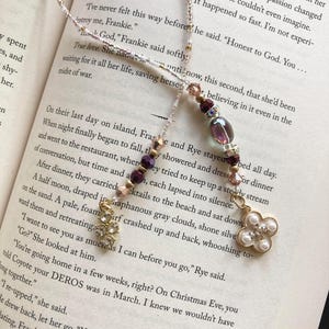 Op de afbeelding: Een close-up van een delicate ketting met een roze en witte kralenketting en een bedel in de vorm van een paarse en gouden bloem met een parel in het midden. De ketting ligt op een boek met tekst over liefde en oorlog.