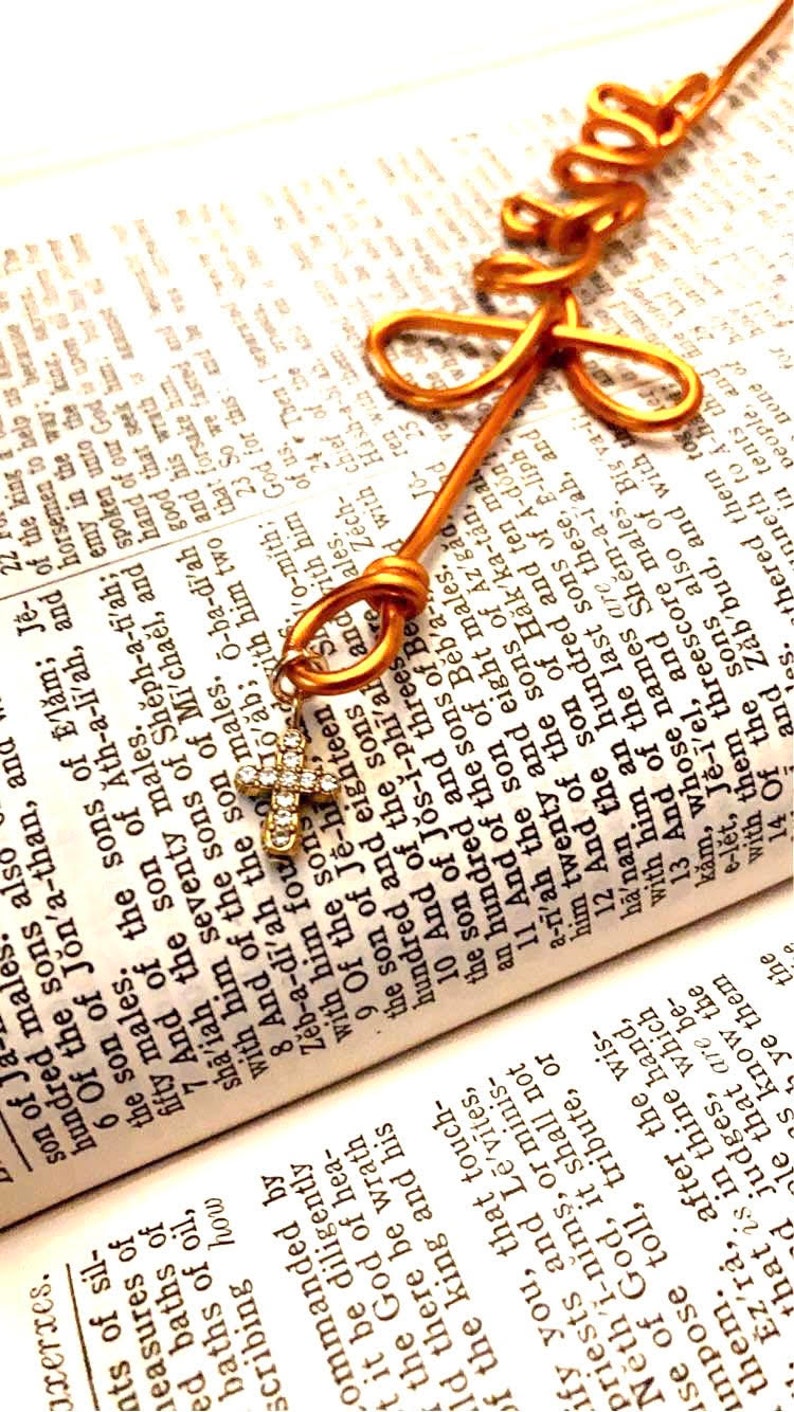 May include: A gold wire bookmark shaped like the word "love" is placed on an open Bible. The bookmark is adorned with a small, sparkling, diamond-encrusted cross.