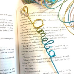 May include: Handmade wire bookmarks in gold, green, and blue. One bookmark features a heart with a small gem, another a stylized letter "O", and the last a spiral design. Colorful wire is coiled in the background.