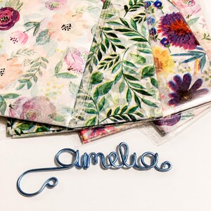 Puede incluir: Una escultura de alambre azul que forma el nombre Amelia en cursiva, colocada frente a varias hojas apiladas de papel translúcido con diversos patrones florales y botánicos en tonos rosa, morado, amarillo y verde.