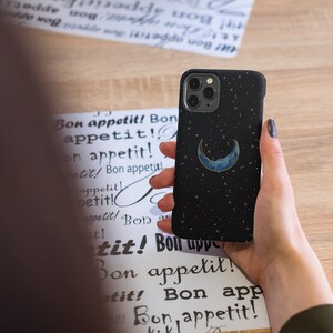 May include: A black phone case with a crescent moon and stars design. The case is made of a soft, textured material and has a subtle shimmer.
