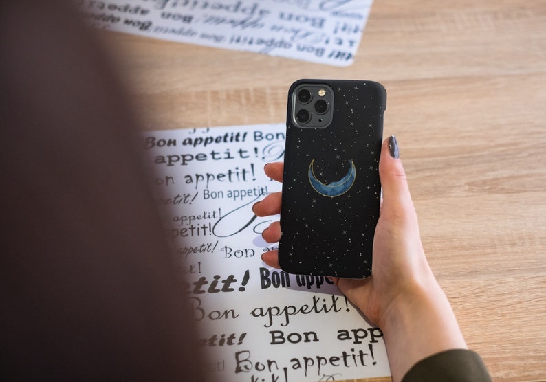 May include: A black phone case with a crescent moon and stars design. The case is made of a soft, textured material and has a subtle shimmer.
