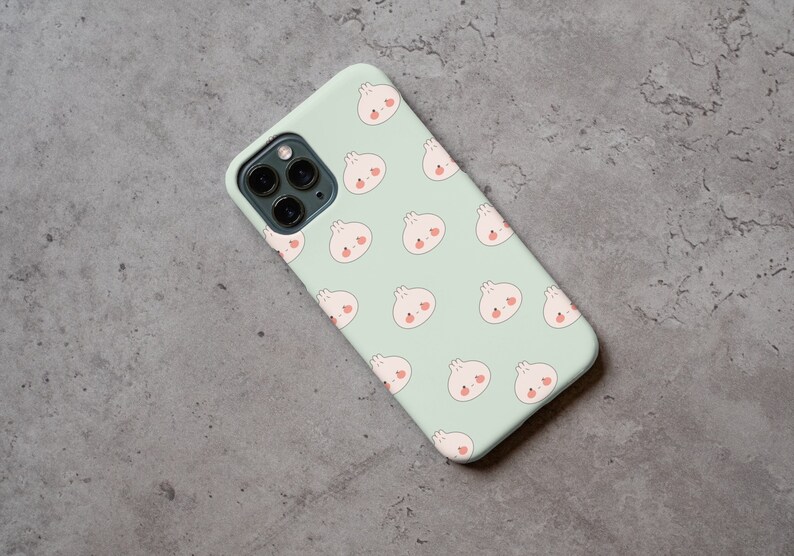 Peut inclure: Une coque de t&eacute;l&eacute;phone vert clair avec un motif r&eacute;p&eacute;t&eacute; de petits pains blancs en dessin anim&eacute; avec des joues roses. La coque a un objectif d'appareil photo noir.