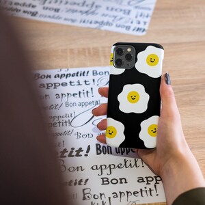 Peut inclure: Une coque de t&eacute;l&eacute;phone noire avec un motif blanc et jaune d'&oelig;ufs au plat souriants. La coque de t&eacute;l&eacute;phone est tenue dans la main d'une personne.