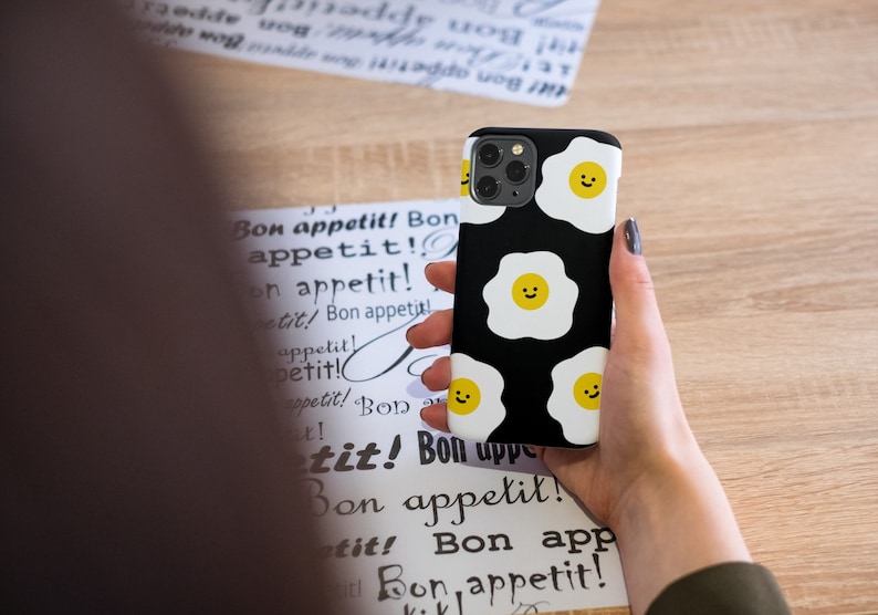 Peut inclure: Une coque de t&eacute;l&eacute;phone noire avec un motif blanc et jaune d'&oelig;ufs au plat souriants. La coque de t&eacute;l&eacute;phone est tenue dans la main d'une personne.