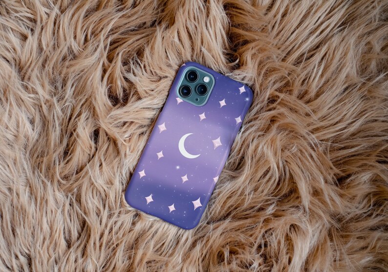 Peut inclure: Une coque de t&eacute;l&eacute;phone violette avec une lune croissante et des &eacute;toiles blanches sur un fond brun moelleux.